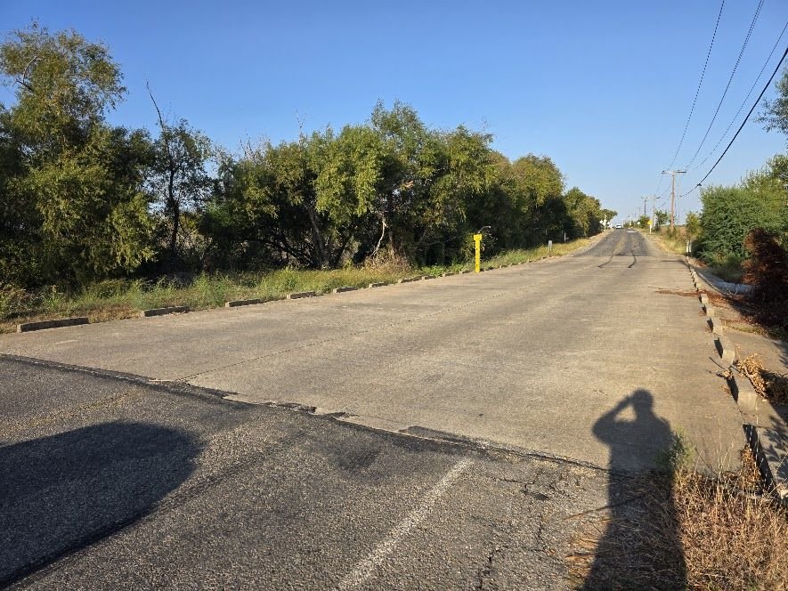 Rural two-lane road with no shoulders or sidewalks and overhead power lines.